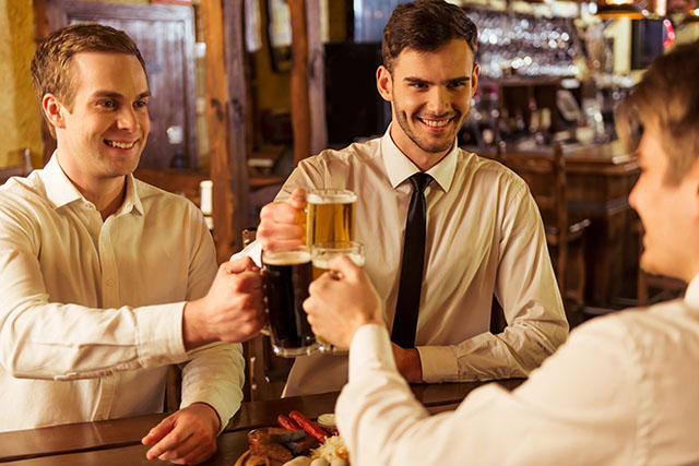 Three young businessmen in white classic shirts are smiling and clanging glasses of beer together while sitting in pub