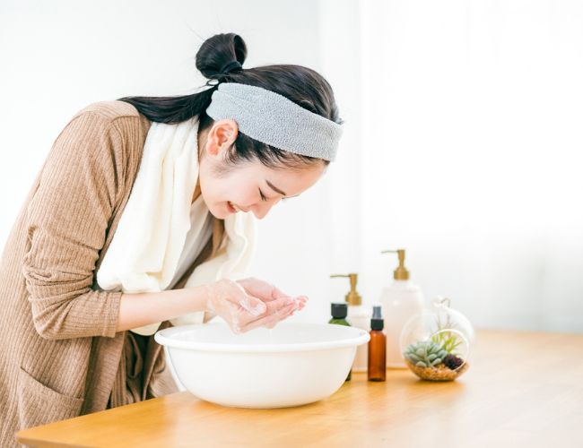 洗面器に溜めた水で顔を洗う女性の写真