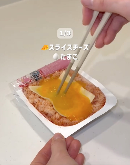 ケチャップとベーコンを混ぜたパックご飯の上にチーズと卵をのせる様子