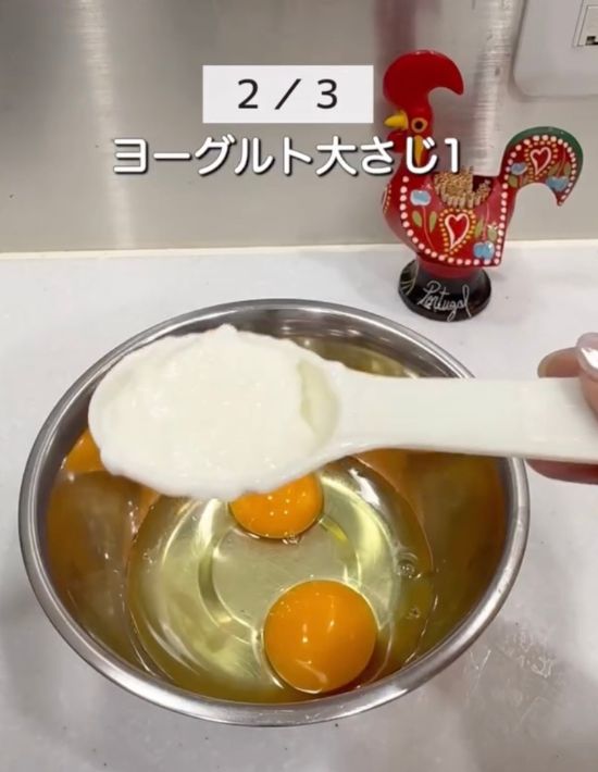 卵を割り入れたボウルを背景に大さじ一杯のヨーグルトを手に持っている様子