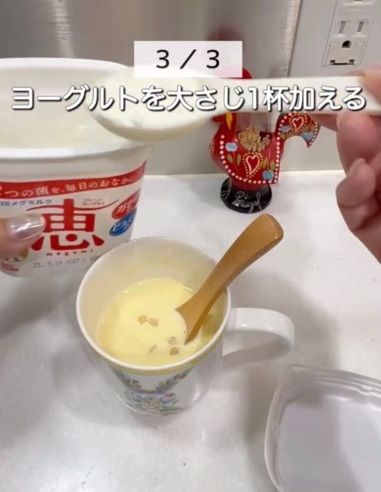 マグカップに入ったコーンスープの上で大さじ１杯のヨーグルトを手に持っている様子