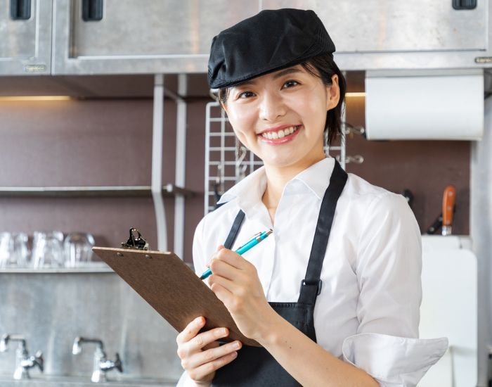帽子とエプロンを着けた女性が笑顔を向けて注文を取る様子